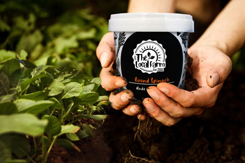 Hands cradling a soil-covered The Local Farm tub of round spinach, in a bed of leafy greens